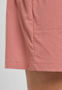 Pantaloni corti di cotone rosa con una texture morbida, caratterizzati da un orlo pulito e un discreto dettaglio della cucitura laterale. Il tessuto sembra leggero e traspirante.