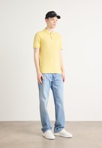 Gul polo-skjorte, lyseblå jeans og hvite joggesko. Svart caps med hvite detaljer. Modellen står på en nøytral bakgrunn.