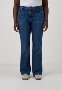 Jeans de mezclilla azul de tiro alto y acampanados, con una textura sutil, diseño de cinco bolsillos y cierre de botón, combinados con zapatillas grises.