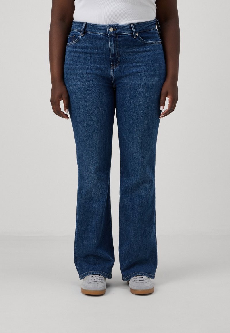 Jeans de mezclilla azul de tiro alto y acampanados, con una textura sutil, diseño de cinco bolsillos y cierre de botón, combinados con zapatillas grises.