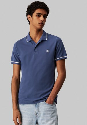Polo de color azul marino con cuello y acentos en las mangas en blanco, que presenta una tapeta de botones y un logo bordado en el lado izquierdo del pecho.