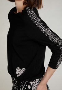 Schwarzer Pullover mit langen Ärmeln, verziert mit weißen Mustern entlang der Schultern und einem Herzdetail am Saum.