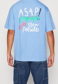 Ljusblå bomulls t-shirt med texten "A.S.A.P! så sakta som möjligt" och en grön alligatorgrafik på baksidan. Kortärmad.