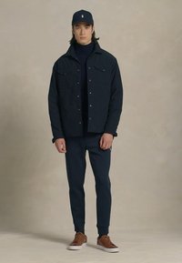 Marineblå jakke og bukser i en avslappet passform, sammen med en marineblå caps og brune sneakers. Jakken har to frontlommer og krage.