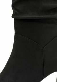 Bottine noire en suède au design épuré, texture lisse, forme incurvée et coutures bien marquées sur les côtés.