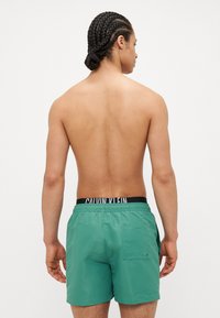 Pantaloni de baie verzi cu un brâu elastic negru, care prezintă logo-ul "Calvin Klein". Materialul pare neted, având un detaliu de buzunar pe spate.