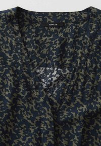 Nahaufnahme eines Kleidungsstücks mit V-Ausschnitt mit dunkelgrünem und marineblauem abstraktem Tarnmuster sowie einem schwarzen Stoffetikett mit der Aufschrift "OPUS" am Halsausschnitt.