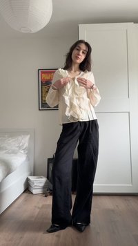 Jauna moteris reguliuoja šviesiai kreminę raukšlėtą palaidinę, dėvi juodas plačias kelnes ir juodus batus, stovi minimalistiniame miegamajame.