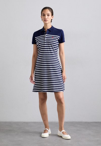 Tummansininen ja valkoinen raidallinen polo-mekko, jossa on lyhyet hihat. Materiaali on puuvillaa. Mekossa on nappilista ja pieni punainen logo rinnassa.