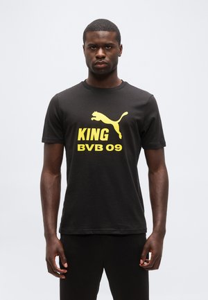 Férfi fekete Puma pólóban, sárga "KING BVB 09" felirattal és Puma logóval, egyszínű világos háttér előtt áll.