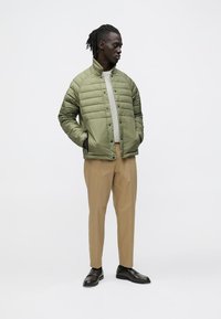 Zelena prešita jakna z visokim ovratnikom, kombinirana s bež hlačami in črnimi čevlji. Ima pritisne gumbe in stranske žepke.