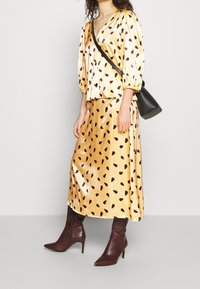 Blouse en satin avec manches bouffantes, col en V, jaune avec motif en goutte marron ; jupe midi assortie ; bottes à talons aigus bordeaux.