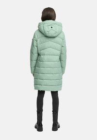 Kvinna som bär en mintgrön vadderad lång vinterjacka med huva, svarta leggings och svarta ankelstövlar, sedd bakifrån.