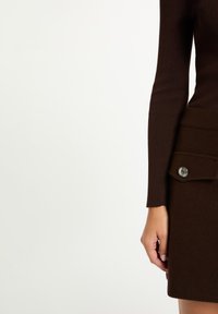 Maglione marrone a maglia con maniche lunghe e vita aderente, abbinato a una gonna marrone scuro con tasca laterale e dettaglio di bottone argentato.