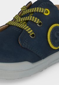 Baskets bleu marine avec lacets en zigzag jaunes et noirs, œillet jaune et détail en patch, et semelle blanche, inclinées pour montrer le côté avant supérieur.