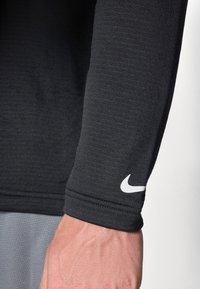 Svart langermet treningsskjorte med strukturert stoff, med en hvit Nike-logo på mansjetten og flate sømmer.