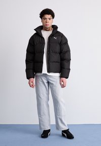 Chaqueta acolchada negra con cuello alto y cierre de cremallera, combinada con jeans de color azul claro y zapatillas negras. Presenta un pequeño logo en el pecho.