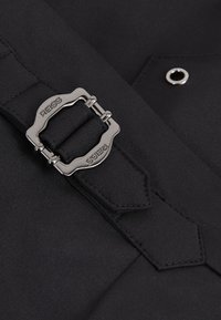 Nagyítva egy fekete szövet öv, rajta téglalap alakú ezüst csat, amelyen a „REISS” felirat látható, egy fekete ruhához rögzítve, melyen egy fém szem is látható.