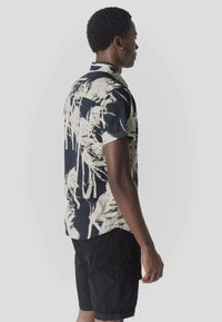 Homme aux cheveux courts et bouclés portant une chemise à manches courtes à imprimé palmiers noir et blanc et un short cargo noir, debout de profil devant un fond uni.