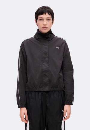 Personne aux cheveux courts et foncés portant une veste de survêtement noire Puma avec des bandes blanches sur les manches et un pantalon noir assorti avec des bandes blanches sur les côtés.