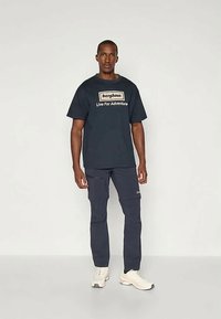 Mann trägt ein navyfarbenes Berghaus-T-Shirt mit dem Aufdruck "Live For Adventure", navyfarbene Cargo-Hosen und weiße Sneakers und steht vor einem einfarbigen Hintergrund.