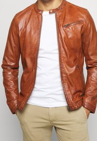 Brun läderjacka med ståkrage, figursydd design, framdragkedja och en dragkedja bröstficka. Bärs över en vit skjorta.