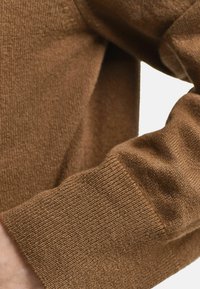 Bruine gebreide trui met een ribgebreide manchette, gemaakt van zachte stof, met een subtiele textuur en een klein merkembleem nabij de manchette.