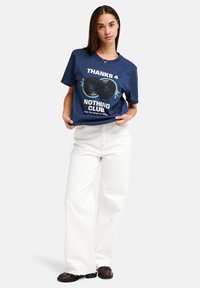 Μπλε ναυτικό βαμβακερό t-shirt με γραφικό που απεικονίζει δύο δίσκους βινυλίου και το κείμενο "THANKS 4 NOTHING CLUB." Συνδυάζεται με λευκό παντελόνι με φαρδιές γραμμές.