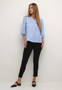 Blouse bleu clair avec de petits motifs en chevrons, manches ballons et coupe décontractée, associée à un pantalon noir ajusté et des mules à talons blocs noires.