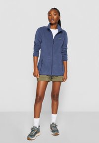 Marine fleece zip-up jakke med høy krage, to sidelommer og en logo på venstre bryst, kombinert med olivengrønne shorts og turløyper.