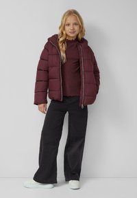 Bordeauxfarbener Pufferjacke mit Kapuze, Reißverschluss und gestepptem Design, getragen über einem burgunderfarbenen, gerippten Oberteil und einer schwarzen, weiten Hose.