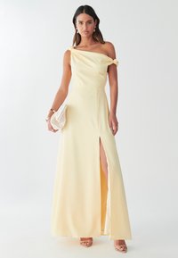 Gul satinkvällsklänning med knutdetalj över axlarna, sidoklyfta och en flödande kjol. Modell som håller i en krämfärgad clutch med textur.