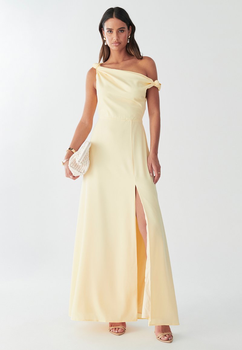 Gul satinkvällsklänning med knutdetalj över axlarna, sidoklyfta och en flödande kjol. Modell som håller i en krämfärgad clutch med textur.