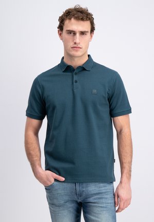 Jonge man met krullend haar die een groenblauw getextureerd poloshirt en lichtblauwe spijkerbroek draagt, staand met één hand in de zak tegen een effen achtergrond.