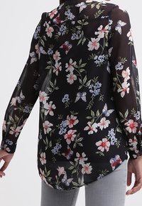 Blouse fleurie en tissu noir transparent, ornée de papillons et de fleurs rouges, roses et bleues. Manches longues, ourlet arrondi et texture douce.