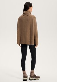 Maglione a collo alto marrone con vestibilità morbida, colletto a costine, spacchi laterali, abbinato a leggings neri e scarpe sportive marroni.
