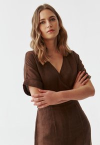 Braunes Leinenkleid mit V-Ausschnitt und kurzen aufgerollten Ärmeln. Der Stoff hat eine sichtbare Textur, und das Design verfügt über eine taillierte Taille und eine gerade Silhouette.
