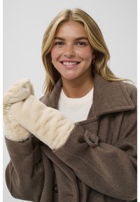 Smiling woman wearing a brown coat and cream-colored faux fur mittens, posing against a plain light gray background.