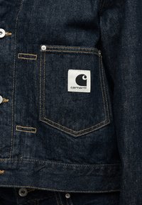 Ποσέτα από σκούρο μπλε τζιν με ορατό μπεζ ράψιμο και λευκή τετράγωνη ετικέτα με το λογότυπο Carhartt σε μαύρο χρώμα.