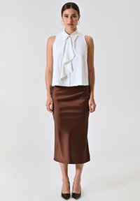 Weißes ärmelloses Oberteil mit Rüschendetail an der Vorderseite, kombiniert mit einem braunen Satin-Midiskirt. Das Model trägt spitze schwarze Absatzschuhe. Schlichte, elegante Silhouette.
