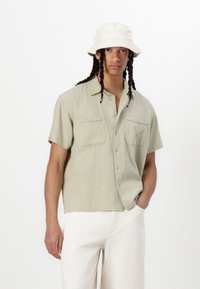 Camicia a maniche corte, di colore verde chiaro, in tessuto texture con due tasche sul petto e chiusura a bottoni, abbinata a pantaloni color crema e un cappello a secchiello.