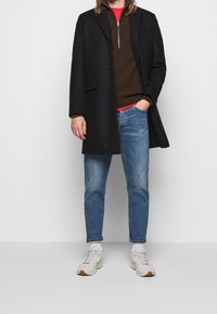 Czarny płaszcz na brązowym swetrze z dzianiny i czerwonej koszuli, w połączeniu z niebieskimi dżinsami i jasnymi butami sportowymi. Prosty design, dopasowany krój.