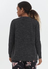 Pull en tricot gris foncé à manches longues, avec un col rond et une finition texturée, arborant une petite étiquette de logo dans l'ourlet.
