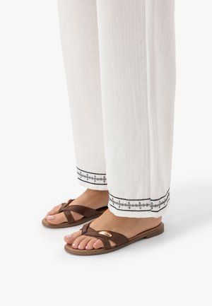 Stopy w brązowych sandałach z paskami krzyżowymi i złotym akcentem, ubrane w białe spodnie z czarnym haftowanym wykończeniem przy kostkach na białym tle.