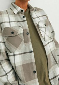 Camicia in flanella a quadri grigia con due tasche sul petto, bottoni neri e una t-shirt verde con scollo rotondo sotto. Tessuto morbido, design casual.