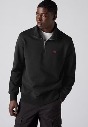 Jeune homme portant un sweatshirt noir à demi-fermeture éclair avec un petit logo rouge, pantalon foncé, debout devant un fond clair uni, regardant vers la gauche.