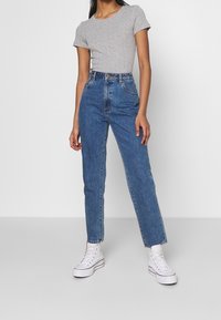 Camiseta ajustada de color gris claro combinada con vaqueros azules de tiro alto y pierna recta, confeccionados en mezclilla, con costuras visibles y bolsillos delanteros. Zapatillas blancas.