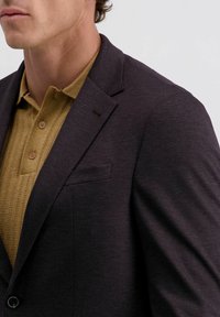 Mann in einem dunkel strukturierten Sakko über einem senfgelben Strickpolohemd mit Knöpfen, von der Mitte der Brust aufwärts vor einem einfarbigen Hintergrund gezeigt.