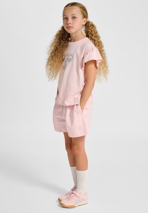 Hellrosa Baumwoll-T-Shirt mit kurzen Rüschensäumen und grafischem Design, kombiniert mit passenden rosa Shorts und hellrosa Sportschuhen.
