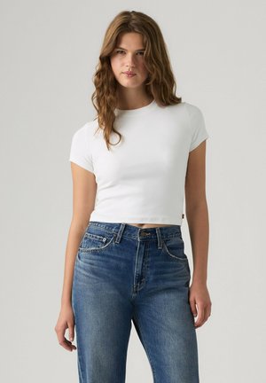 Młoda kobieta z falowanymi brązowymi włosami, ubrana w dopasowany biały krótki T-shirt i niebieskie jeansy z wysokim stanem na jednolitym jasnym tle.